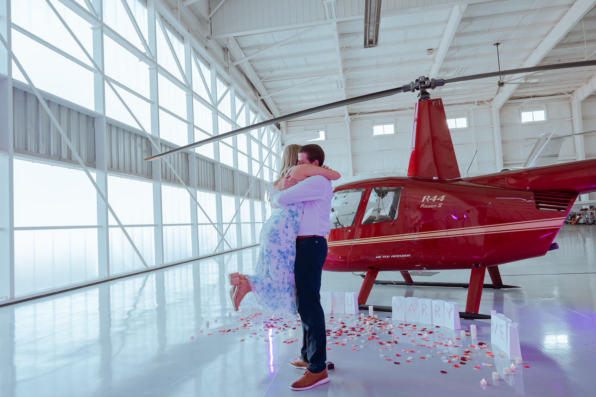 Couple embracing in a hangar beside a red helicopter with a Will You Marry Me display